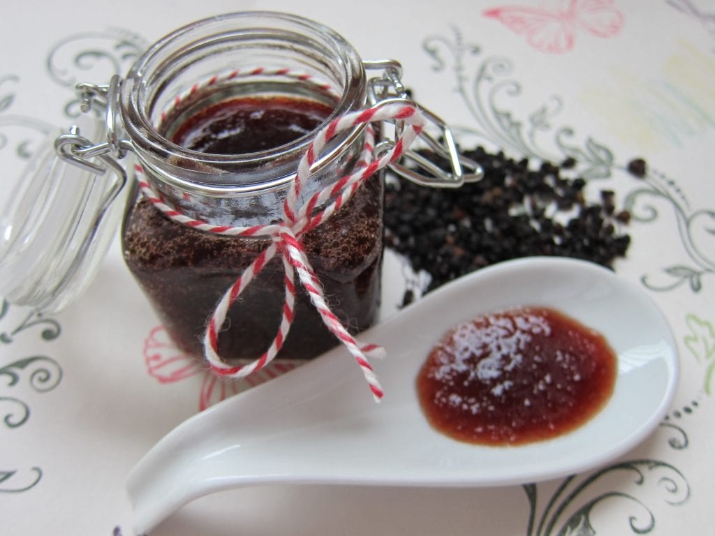 A jar of homemade jam tied with red and white twine, next to a spoonful of jam and black peppercorns on a floral tablecloth.